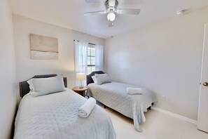 Quarto com duas camas, ventilador de teto, janela com cortinas e porta.