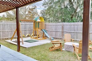 A backyard with a wooden swing set, a slide, and two Adirondack chairs.