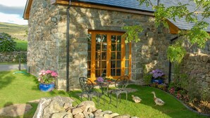 Una casa de piedra con puerta de madera, un pequeño patio con mesa y sillas, y un jardín con flores y un pato.