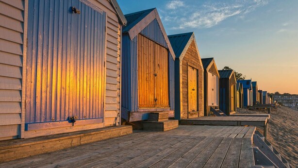 Une rangée de cabanes de plage en bois, dotées de toits inclinés, est disposée le long d’une promenade en bois.