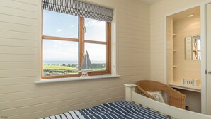 Un dormitorio con un ventanal grande, una cama, una silla de mimbre y una pequeña estantería con objetos.