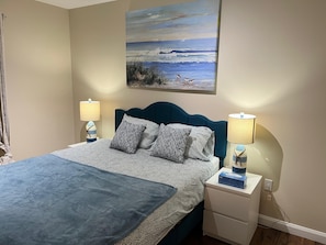 A neatly made bed with patterned bedding, two bedside lamps, a nightstand, and a painting of a seascape above the bed.