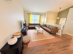 A living room with a brown leather sofa, a coffee table, and a TV stand. There is a window with yellow curtains and a white railing.