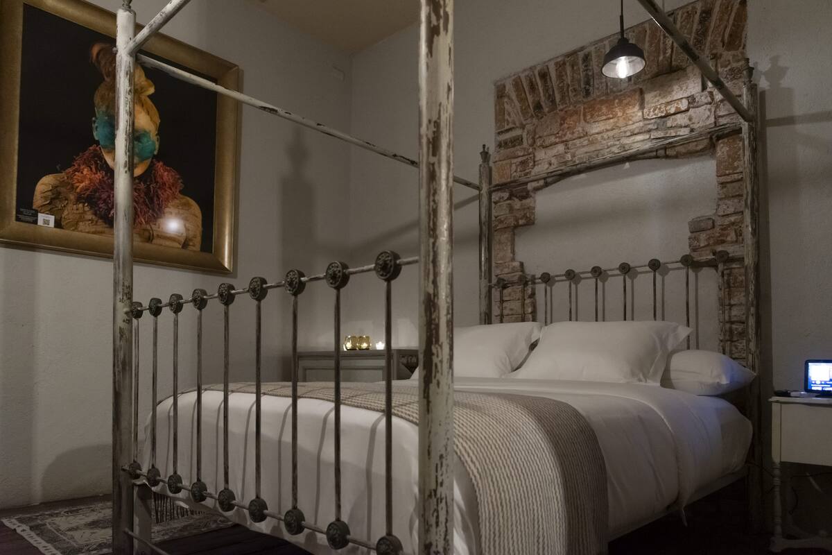 Un dormitorio con estructura de cama de metal, pared de ladrillo, cabecera de madera, mesita de noche con lámpara y un cuadro en la pared.