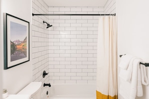 Un baño con azulejos blancos tipo metro, un cuadro en la pared, una bañera y una cortina de ducha.