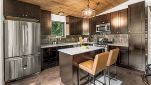 A modern kitchen with dark wood cabinets, stainless steel appliances, a central island with bar stools, and a wooden ceiling. - NEW Hot Tub + Arcade + Fire Pit + Mtn Views (Bailey)