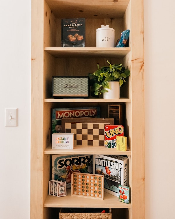 Un estante de madera con varios juegos de mesa, un libro titulado 'The Camp & Cabin Cookbook', un recipiente blanco con la etiqueta 'WOOF' y una planta en maceta.