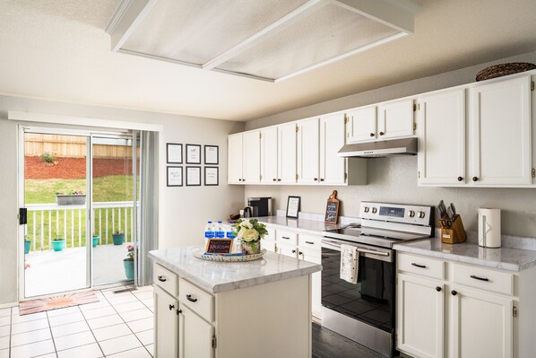 Cocina con armarios blancos, una isla central, estufa y una ventana grande con vistas a un jardín.