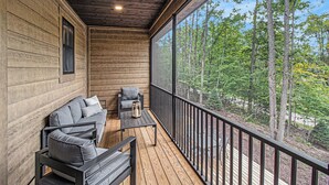 Una terraza de madera con un sofá y sillones, una mesa y un ventanal grande con vistas a un bosque.