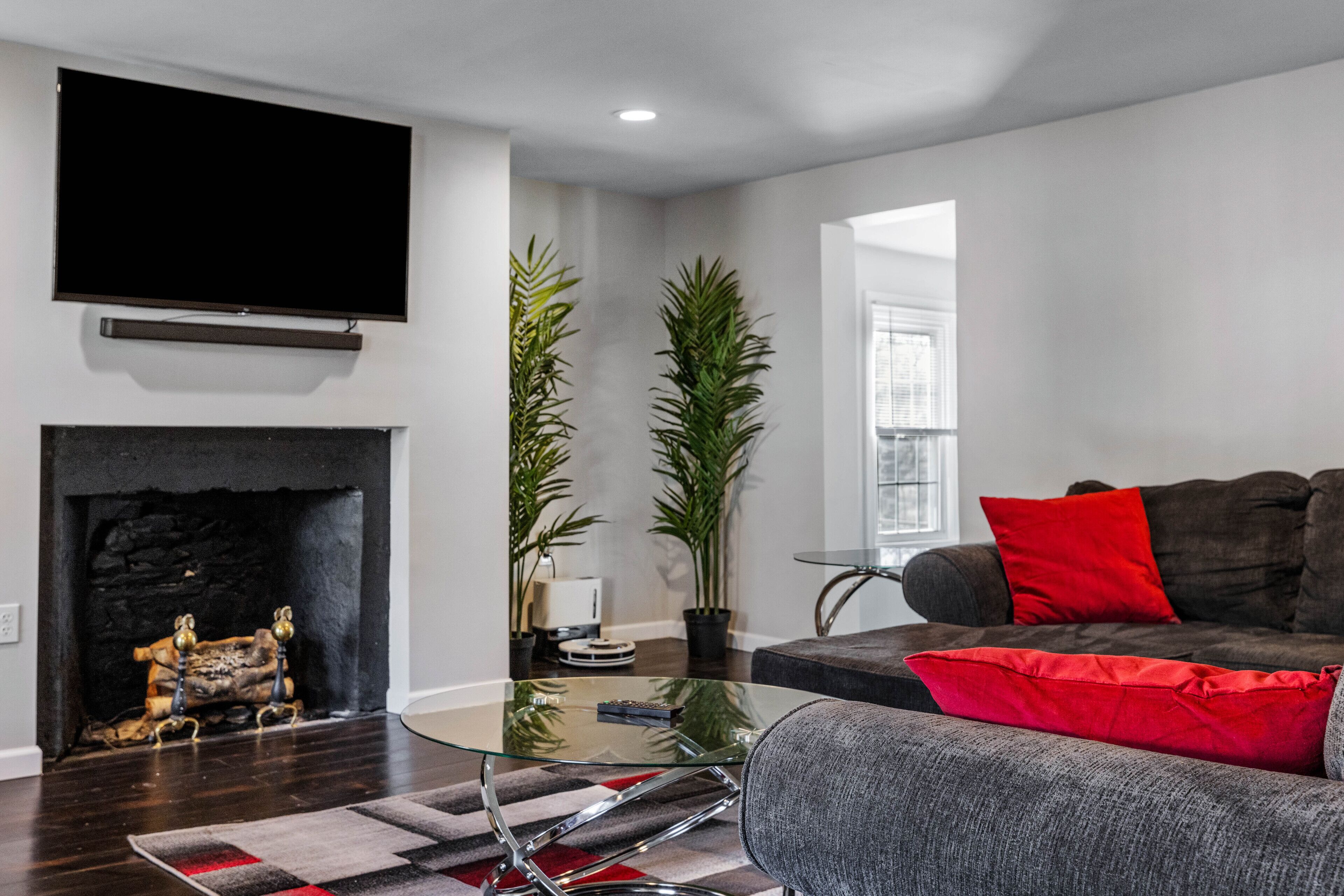 A modern living room with a fireplace, a flat-screen TV mounted above it, a grey sofa with red cushions, and two tall potted plants.