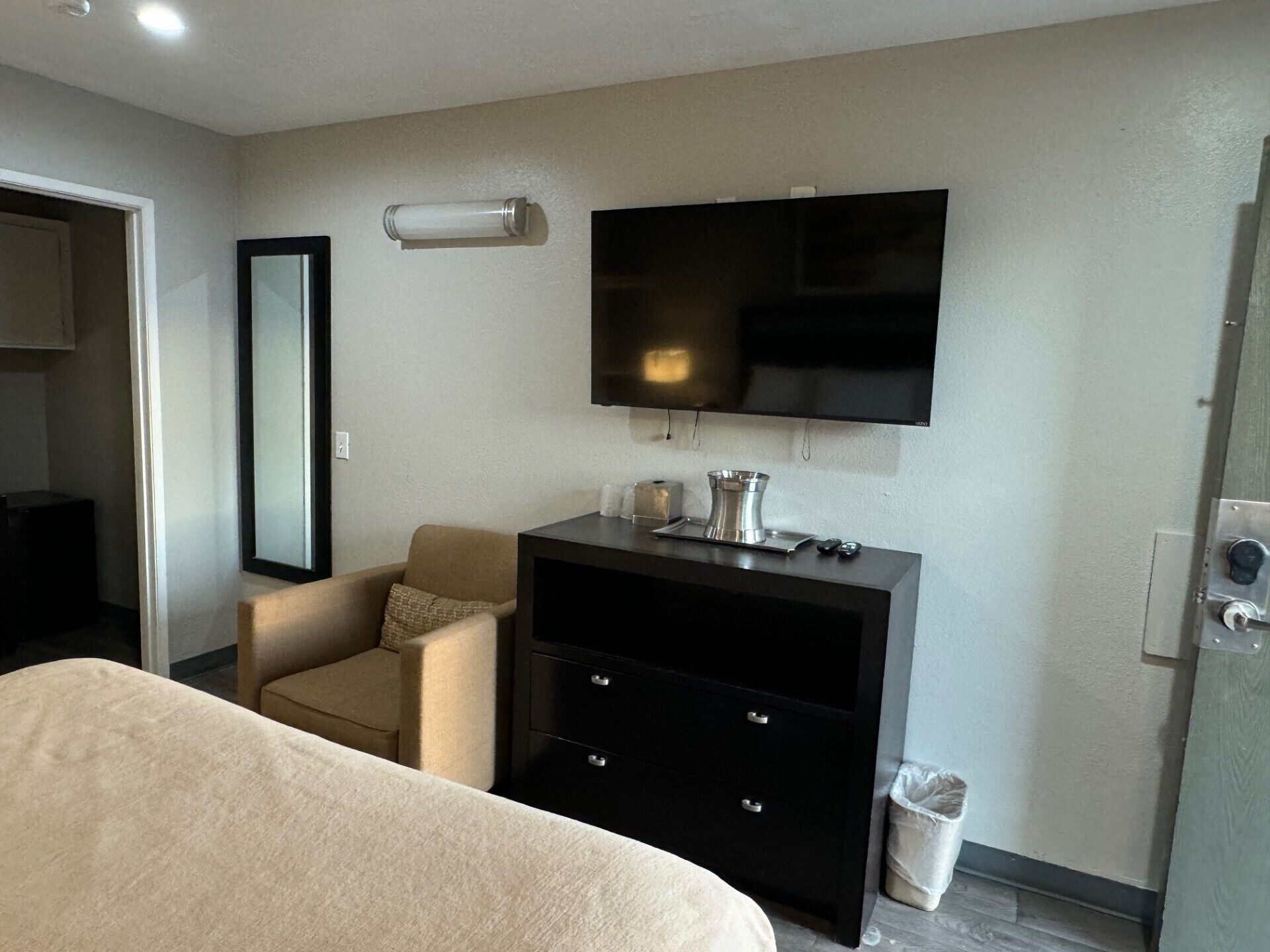 A hotel room with a bed, a TV mounted on the wall, a small desk with drawers, a chair, and a trash bin.