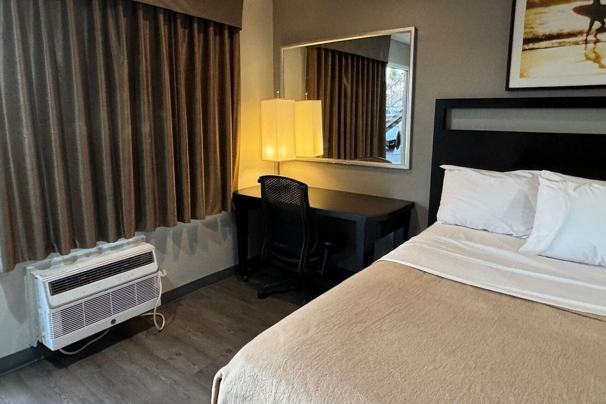 Habitación de hotel con cama, escritorio con silla, lámpara, espejo grande y unidad de aire acondicionado.