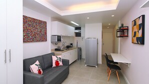 A compact kitchen with white cabinets, a grey sofa with floral cushions, and a small dining area with a white table and black chairs. - Rio Spot Homes Copacabana C059 (Rio de Janeiro)