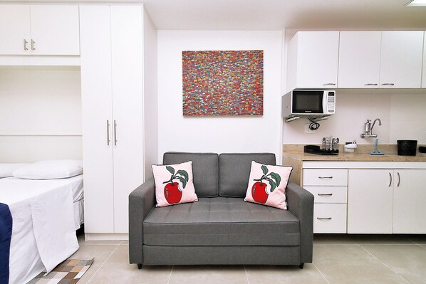 A compact living space with a grey sofa, white cabinets, a microwave, and a colorful abstract painting.