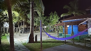 A nighttime view of a residential area with a hammock, palm trees, a blue house, and parked cars. - Pousada Costa Azul (Maraú)
