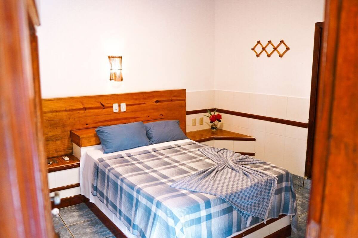 Une chambre à coucher avec une tête de lit en bois, un lit recouvert d’une couverture à carreaux bleus et blancs, une table de chevet avec une lampe et une petite étagère ornée d’un vase de fleurs.
