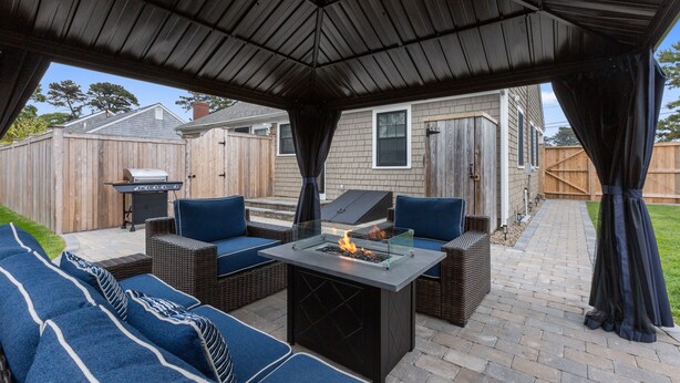 Une terrasse extérieure dotée d’un foyer à gaz avec un dessus en verre, de sièges rembourrés bleus et d’une pergola au-dessus.