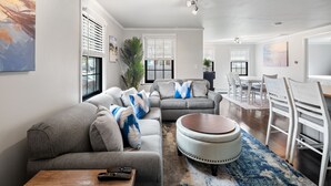 Un salon moderne avec un canapé d’angle gris, un pouf rond et une table basse en bois. On y trouve également un coin repas avec une table et des chaises blanches.
