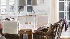 Een gezellig eetgedeelte met rieten stoelen en een houten tafel, een keuken op de achtergrond met witte kasten, en een groot raam dat natuurlijk licht binnenlaat.