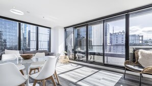 A modern living room with a glass table, white chairs, a sofa, and a large window offering a city view. - Riverview Suites by EdgeTrip (Melbourne)