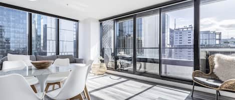 A modern living room with a glass table, white chairs, a sofa, and a large window offering a city view.