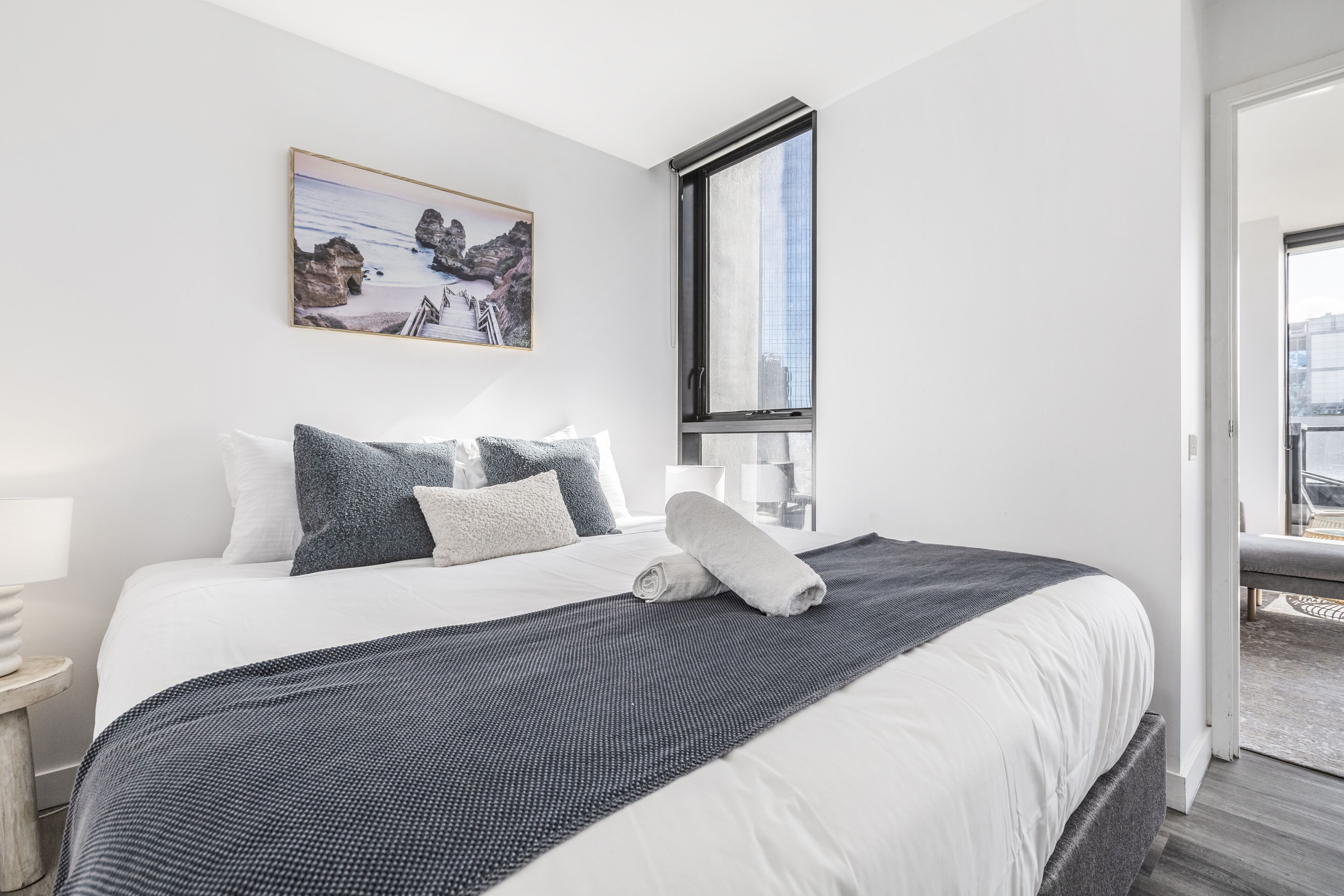 A neatly made bed with a view of the city through a large window.