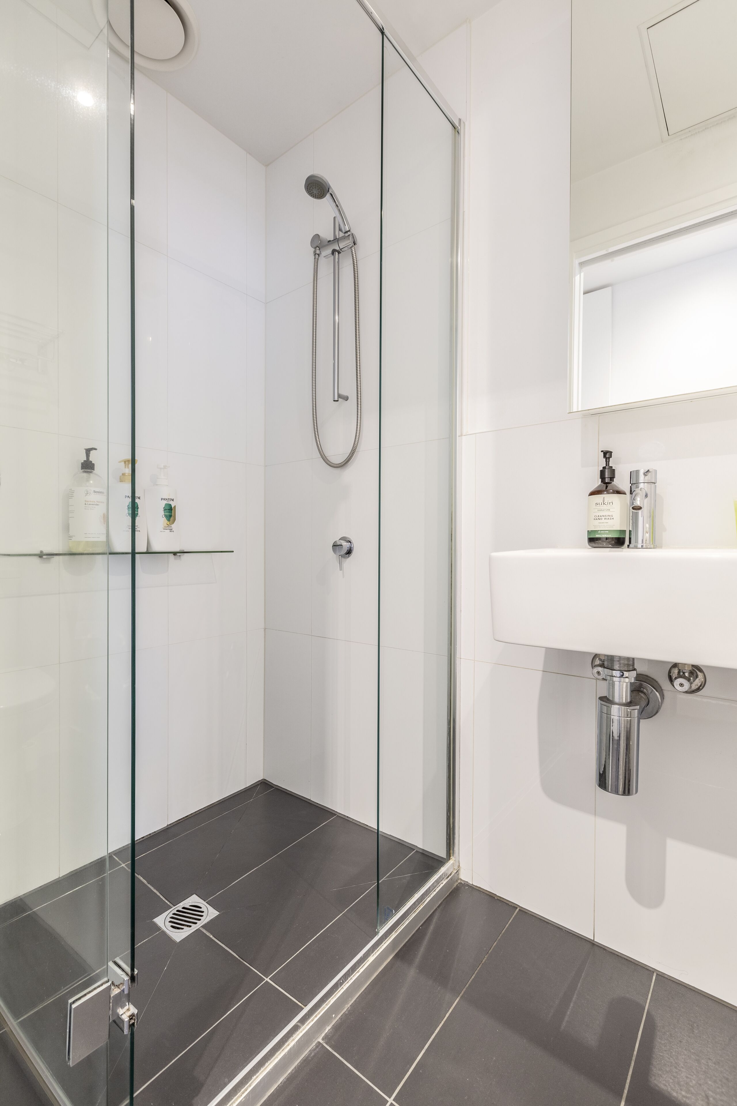 Ein modernes Badezimmer mit einer Duschkabine aus Glas, einem weißen Waschbecken mit Spiegel darüber und einem Ablagebereich für Toilettenartikel.