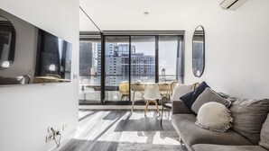 A modern living room with a sofa, a dining table, and chairs. There is a large window with a view of buildings. - Riverview Suites by EdgeTrip (Melbourne)