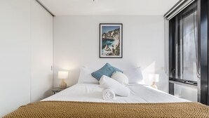 A neatly made bed with white linens, a blue throw pillow, and a rolled towel. There is a framed picture of a coastal landscape on the wall above the bed. - Riverview Suites by EdgeTrip (Melbourne)