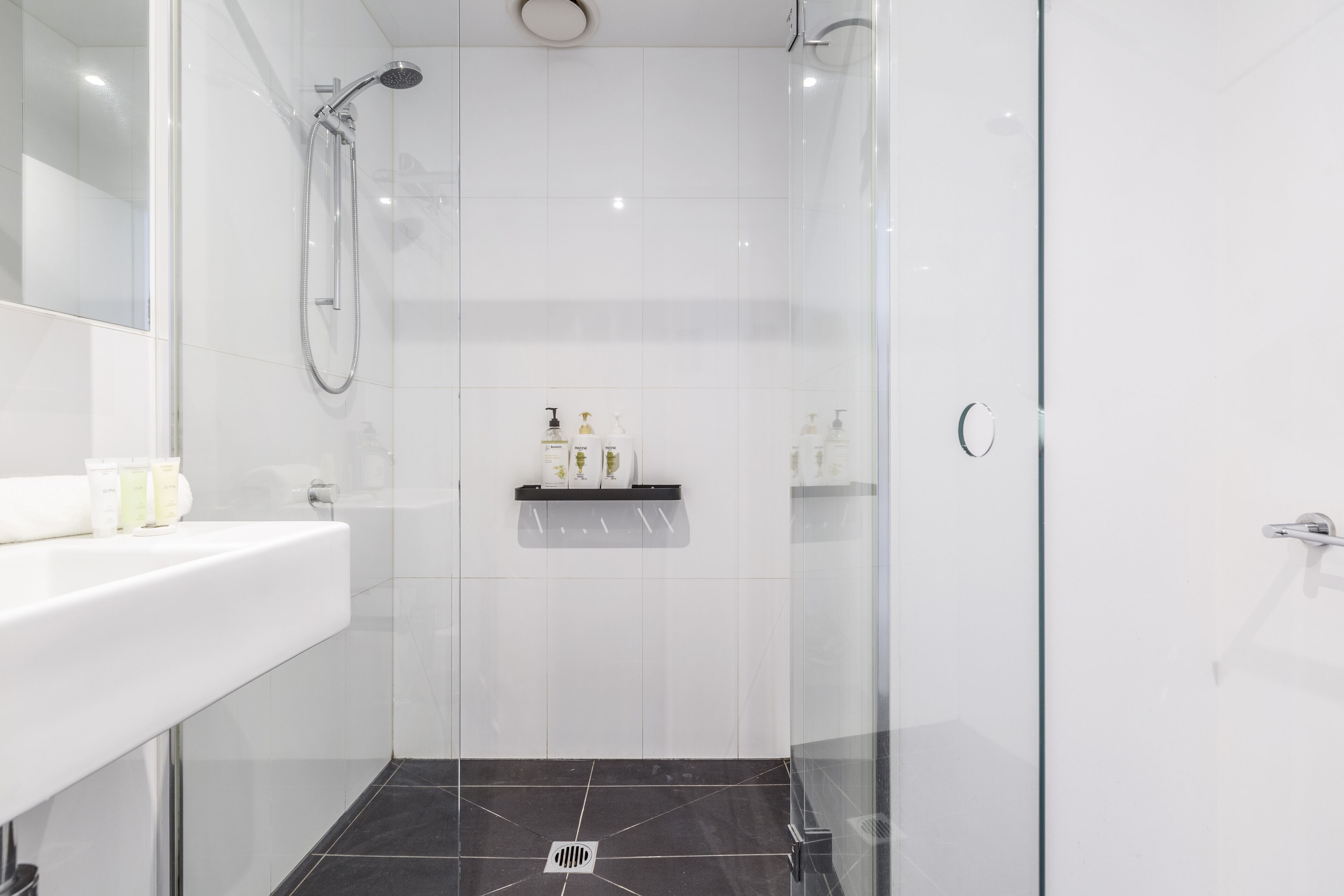 A modern bathroom with a glass shower enclosure, a white sink with toiletries, and a black tiled floor.