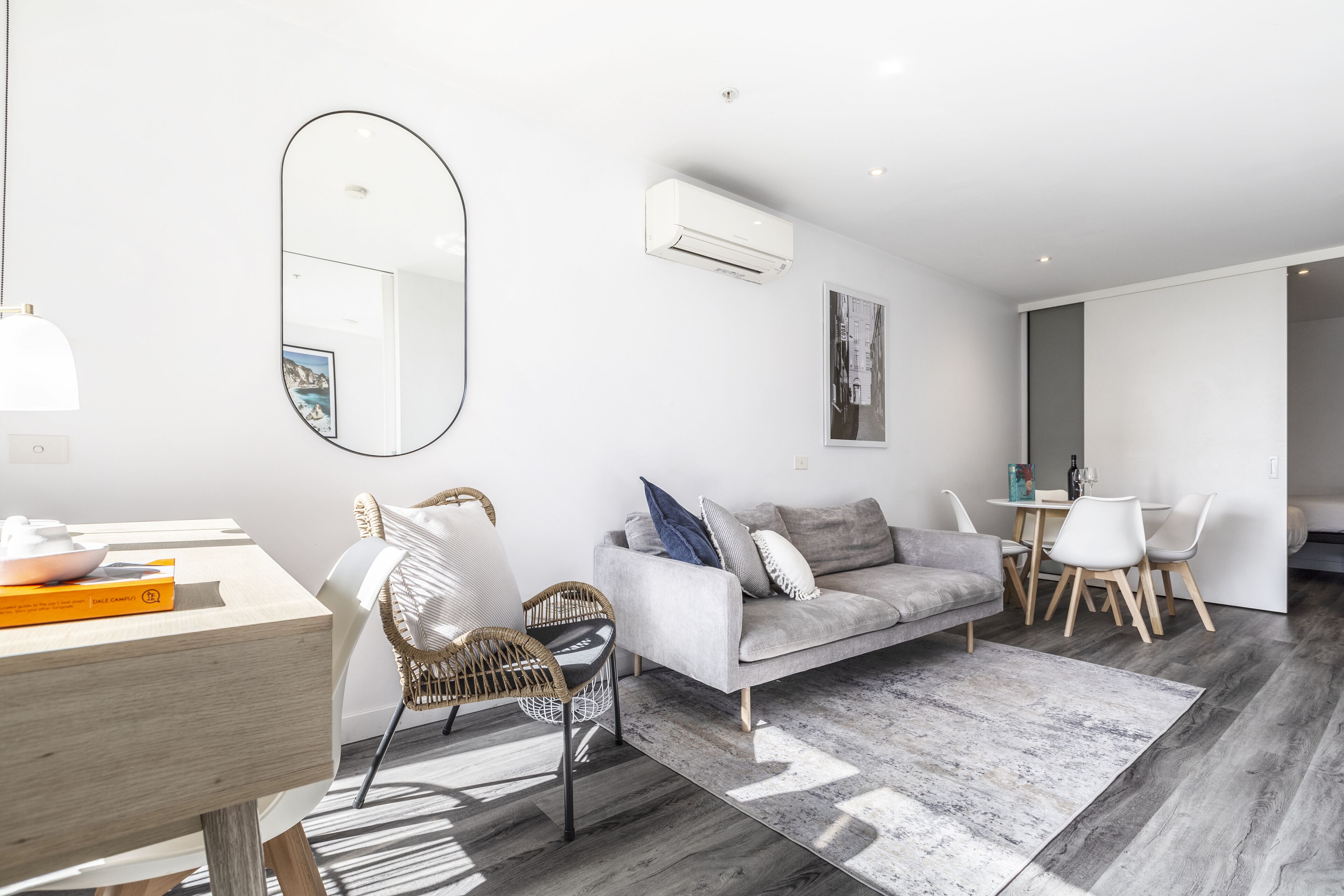 A modern living room with a grey sofa, wicker chairs, a dining table with chairs, and a large mirror.