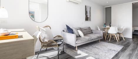 A modern living room with a grey sofa, wicker chairs, a dining table with chairs, and a large mirror.