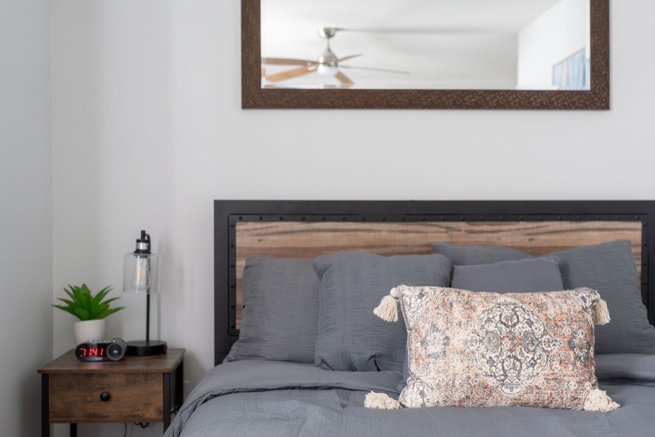A bedroom with a wooden headboard, grey bedding, a decorative pillow, a bedside table with a plant, a lamp, and a digital clock.