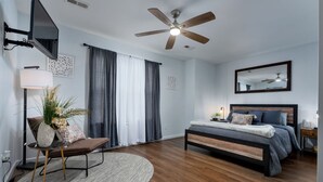 Un dormitorio con cama, escritorio, silla, alfombra redonda, ventilador de techo y televisor montado en la pared.