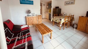 A compact living space with a sofa, a wooden table, a dining table with chairs, a kitchen area with a refrigerator, and a ceiling fan. - Nice 2 room with cabin swimming pool Richelieu Cap d'Agde (LE CAP D AGDE)