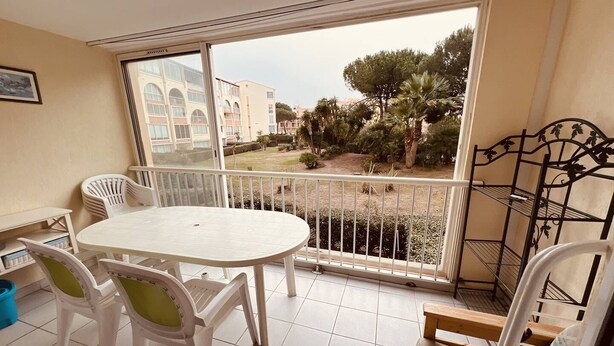 A balcony with a white table and chairs, a metal railing, and a view of buildings and trees. - Nice 2 room with cabin swimming pool Richelieu Cap d'Agde (LE CAP D AGDE)