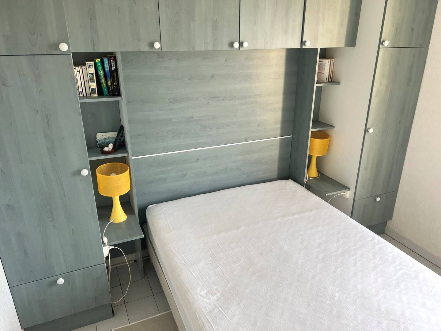 A bedroom with a bed, a bedside table, a lamp, and built-in shelving with books and decorative items.