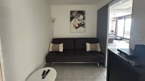 A modern living room with a dark sofa, a painting on the wall, a small white table, and a view through a glass door. - Pretty 3 Room Apartment with 2 alcoves, AIRCON and WIFI, sea view, pool Rochelongue Cap d'Agde (LE CAP D AGDE)