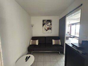 Un salon moderne avec un canapé foncé, un tableau accroché au mur, une petite table blanche et une vue à travers une porte vitrée.