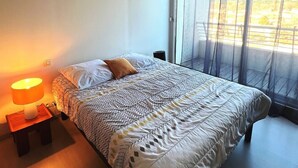 A modern bedroom with a large bed, a bedside lamp, and a view through a window with sheer curtains. - Pretty 3 Room Apartment with 2 alcoves, AIRCON and WIFI, sea view, pool Rochelongue Cap d'Agde (LE CAP D AGDE)