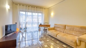 A living room with a sofa, a TV, a wooden table, and a large window with sheer curtains.