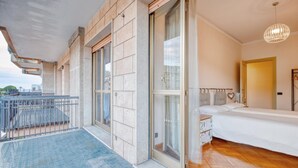 A bedroom with a bed, a bedside table, and a window. There is a balcony with a railing and a view of the outside.