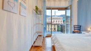 A bedroom with a bed, a chair, a table, a plant, and a view of buildings through a balcony door.