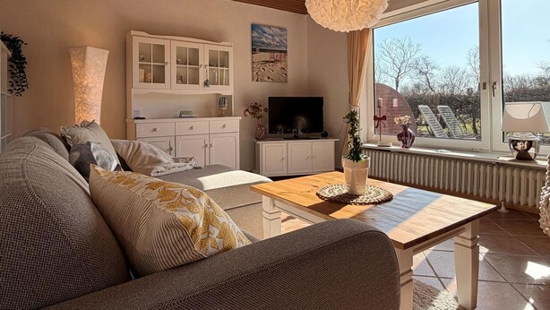 Ein Wohnzimmer mit Holzbalkendecke, einer Couch, einem Couchtisch, einem Fernseher und einem Fenster mit Blick ins Freie.