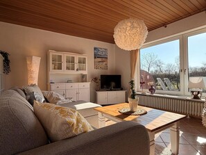 A living room with a wooden ceiling, a sofa, a coffee table, a TV, and a window with a view of the outdoors. - Haus Ruhewinkel 2 - Bungalow Ruhewinkel 2 (Alkersum)