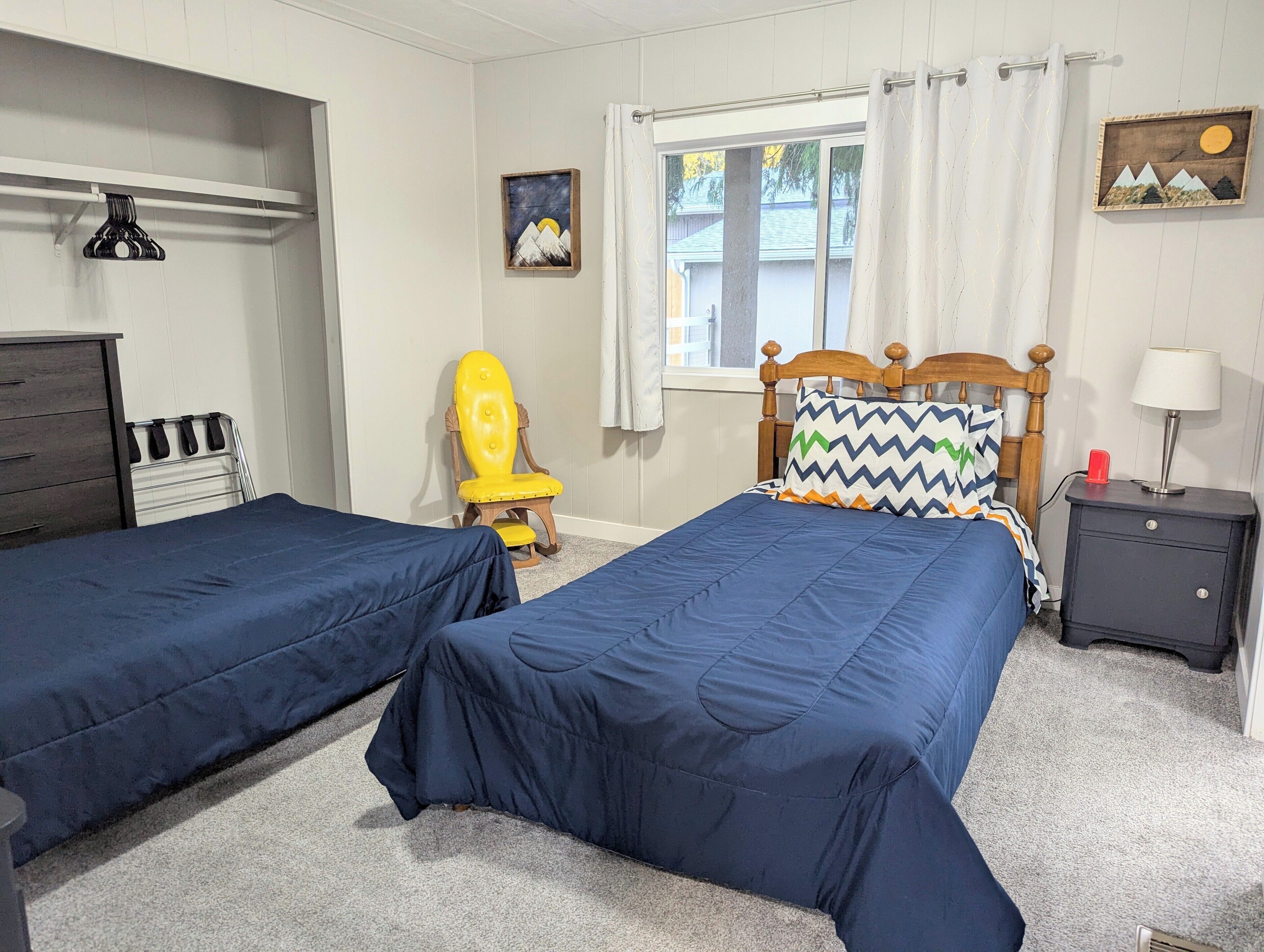 A bedroom with two single beds, a wooden chair, a nightstand, a dresser, and a window with curtains.