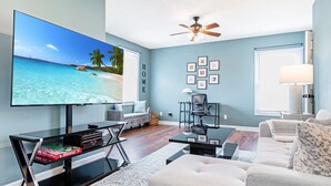 A living room with a large flat-screen TV displaying a beach scene, a ceiling fan, a sofa, a coffee table, and a wall with framed pictures. - Cozy pet-friendly home with fenced backyard, fully equipped for long-term stays. (Louisville)