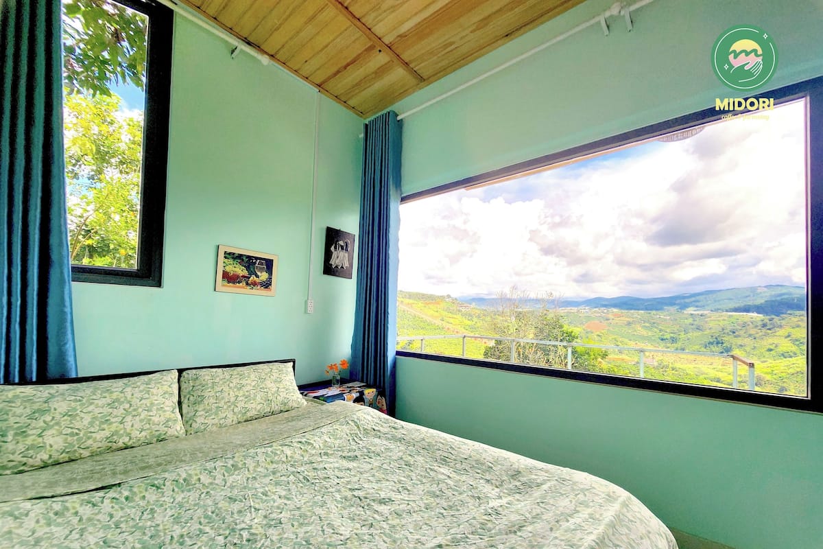 A bedroom with a large window, a bed, and a view of a green landscape.