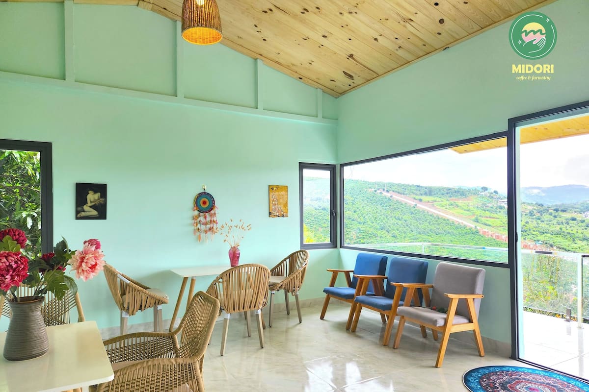 A spacious room with a large window offering a view of a green landscape. There are wicker chairs and a table with a vase of flowers.