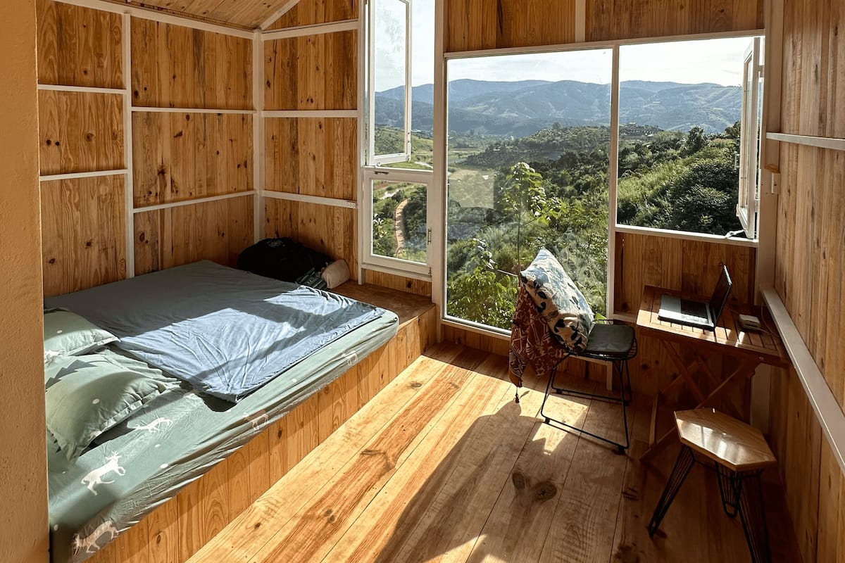 A wooden room with a bed, a desk, and a large window offering a view of the countryside.